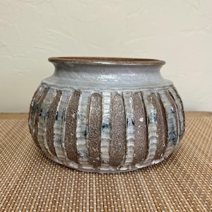 May include: A handcrafted ceramic vase with a wide, rounded body and a textured, ribbed exterior. The vase features a light gray upper section and a brown and white lower section. The interior is a darker brown. The vase is sitting on a bamboo mat.