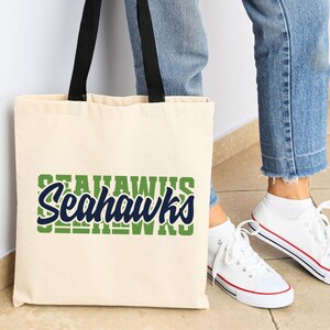 Puede incluir: Un bolso tote de color crema con asas negras que muestra la palabra "Seahawks" en verde y azul marino. El bolso se muestra junto a una persona que lleva vaqueros azules y zapatillas blancas.
