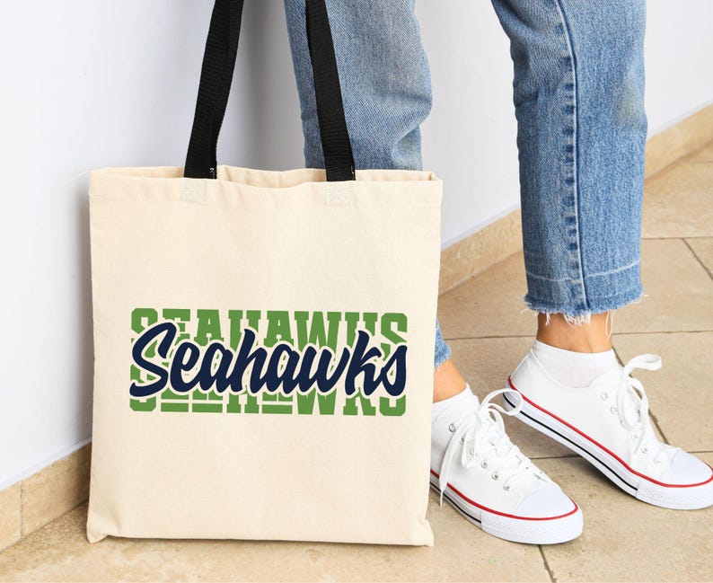 Puede incluir: Un bolso tote de color crema con asas negras que muestra la palabra "Seahawks" en verde y azul marino. El bolso se muestra junto a una persona que lleva vaqueros azules y zapatillas blancas.
