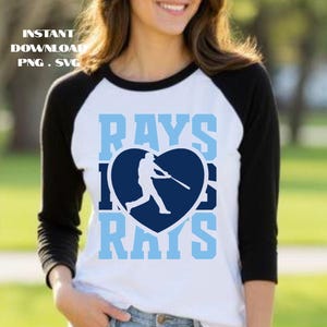 May include: A white baseball raglan shirt with black sleeves. The shirt features the word "RAYS" in light blue, with a baseball player silhouette inside a navy blue heart.