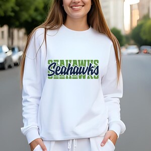 Puede incluir: Sudadera blanca con la palabra "Seahawks" en verde y azul. La sudadera tiene mangas largas y cuello redondo. El texto est&aacute; apilado, con "Seahawks" en una fuente audaz y estilizada.
