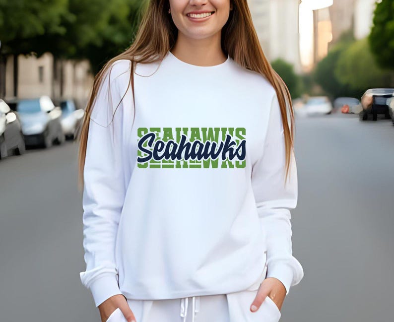 Puede incluir: Sudadera blanca con la palabra "Seahawks" en verde y azul. La sudadera tiene mangas largas y cuello redondo. El texto est&aacute; apilado, con "Seahawks" en una fuente audaz y estilizada.