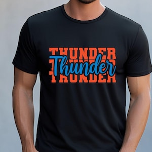 May include: Black t-shirt with the word "Thunder" in orange block letters stacked three times. The word "Thunder" is written in a cursive blue font in the middle of the orange block letters.