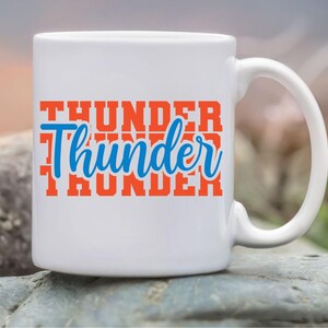 May include: White ceramic mug with the word "Thunder" in orange block letters and a blue cursive "Thunder" overlay. The mug has a curved handle and is set against a blurred outdoor background.