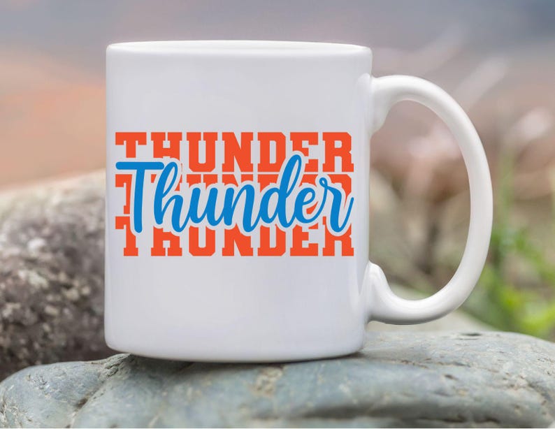 May include: White ceramic mug with the word "Thunder" in orange block letters and a blue cursive "Thunder" overlay. The mug has a curved handle and is set against a blurred outdoor background.