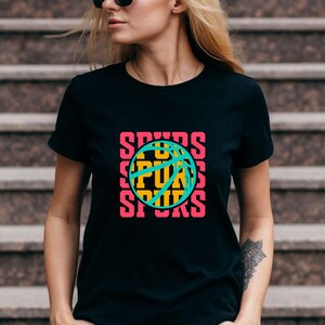 May include: Black t-shirt with a basketball graphic. The word "SPURS" is stacked vertically on the left and right sides of the basketball. The basketball is teal with yellow and pink accents. The shirt is short-sleeved.