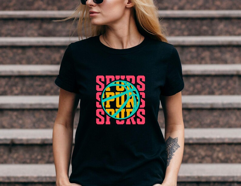 May include: Black t-shirt with a basketball graphic. The word "SPURS" is stacked vertically on the left and right sides of the basketball. The basketball is teal with yellow and pink accents. The shirt is short-sleeved.