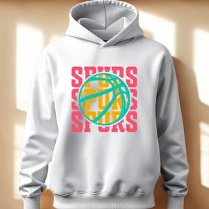 May include: White hoodie with a drawstring hood. The front features a basketball graphic with the word "SPURS" in pink, teal, and yellow. The design is repeated three times vertically. A casual, sporty apparel item.