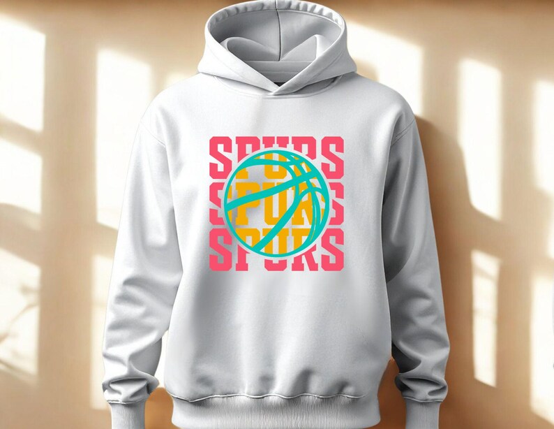 May include: White hoodie with a drawstring hood. The front features a basketball graphic with the word "SPURS" in pink, teal, and yellow. The design is repeated three times vertically. A casual, sporty apparel item.