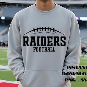 May include: A light gray sweatshirt with a black graphic of a football and the words "RAIDERS FOOTBALL". The sweatshirt also has the words "INSTANT DOWNLOAD PNG . SVG" in black text. The person is standing on a football field.