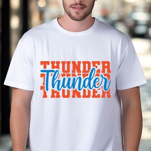 May include: White t-shirt with the word "Thunder" in a blue script font, layered over the word "Thunder" in a bold orange font. The shirt is a crew neck style.