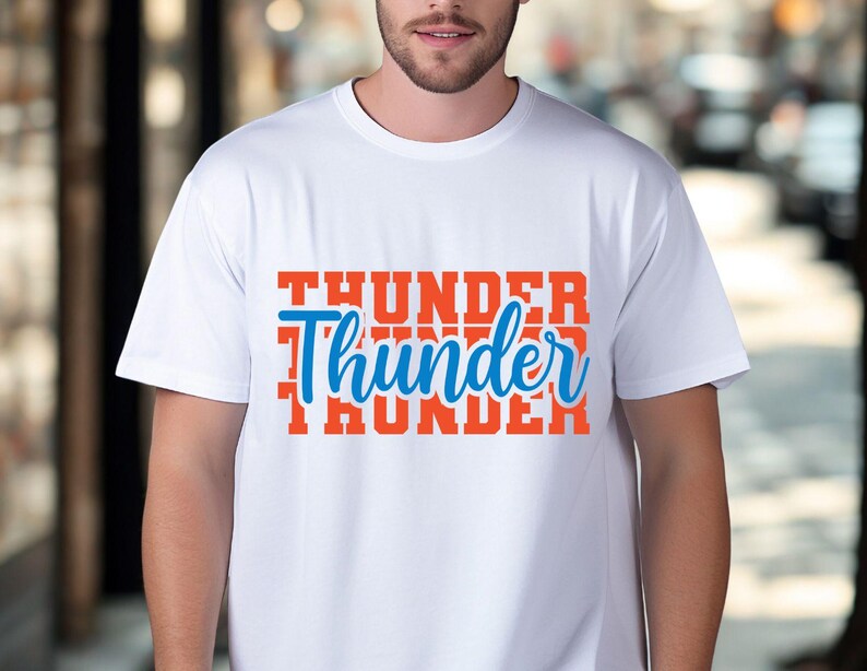 May include: White t-shirt with the word "Thunder" in a blue script font, layered over the word "Thunder" in a bold orange font. The shirt is a crew neck style.