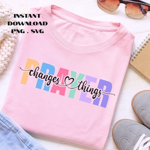 Peut inclure: T-shirt rose clair avec l'inscription "PRAYER changes things" en lettres colorées et cursives. Le t-shirt est présenté avec des lunettes de soleil, un carnet et des baskets grises.