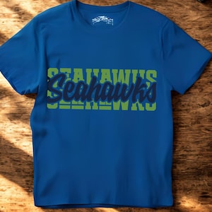 Puede incluir: Una camiseta azul con la palabra "Seahawks" en un dise&ntilde;o verde y azul en la parte delantera. La camiseta de manga corta est&aacute; hecha de un material suave. La camiseta se muestra sobre una superficie de madera.