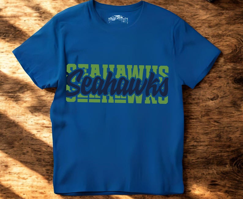 Puede incluir: Una camiseta azul con la palabra "Seahawks" en un dise&ntilde;o verde y azul en la parte delantera. La camiseta de manga corta est&aacute; hecha de un material suave. La camiseta se muestra sobre una superficie de madera.
