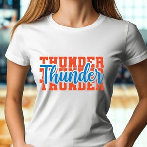 May include: White t-shirt with the word "Thunder" in orange block letters stacked vertically, with the word "Thunder" in blue script in the center. The shirt is short-sleeved and appears to be made of cotton.