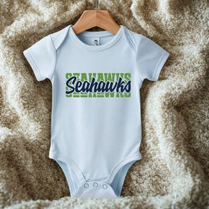 Puede incluir: Un body de beb&eacute; azul claro con el texto "Seahawks" en verde y azul marino. El body est&aacute; colgado de una percha de madera sobre un fondo texturizado de color crema.