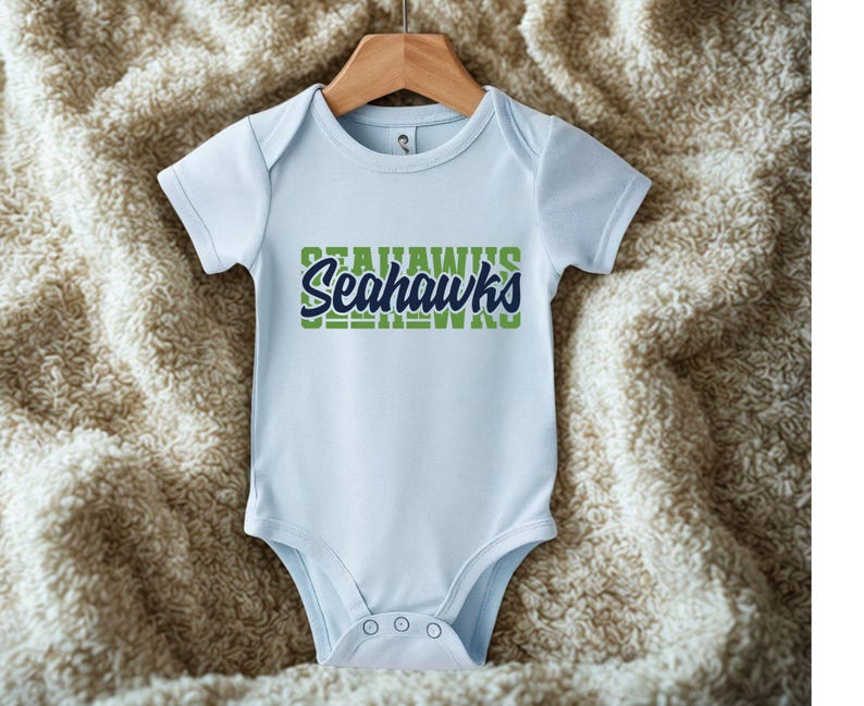 Puede incluir: Un body de beb&eacute; azul claro con el texto "Seahawks" en verde y azul marino. El body est&aacute; colgado de una percha de madera sobre un fondo texturizado de color crema.