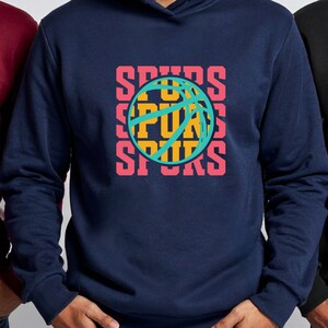 May include: A navy blue hoodie with a basketball graphic. The graphic features the word "SPURS" in a stacked design with a basketball in the center. The text and basketball details are in shades of pink, yellow, and teal.