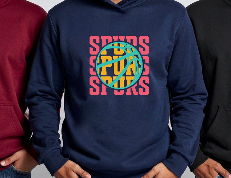 May include: A navy blue hoodie with a basketball graphic. The graphic features the word "SPURS" in a stacked design with a basketball in the center. The text and basketball details are in shades of pink, yellow, and teal.