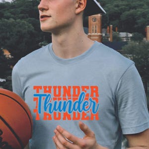 May include: A light blue t-shirt with the word "Thunder" in orange and blue. A basketball with the Wilson logo is in the foreground. The shirt is a crew neck style.