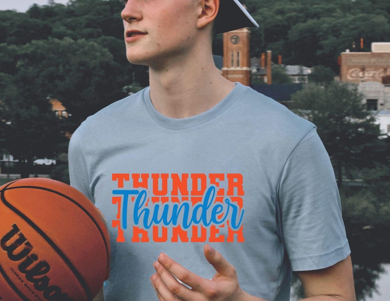 May include: A light blue t-shirt with the word "Thunder" in orange and blue. A basketball with the Wilson logo is in the foreground. The shirt is a crew neck style.
