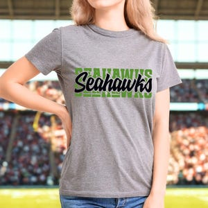 Puede incluir: Una camiseta gris con la palabra "Seahawks" en letras verdes y negras. La camiseta es de manga corta y parece estar hecha de un material suave. El fondo muestra una imagen borrosa de un estadio.