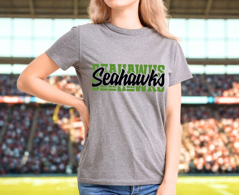 Puede incluir: Una camiseta gris con la palabra "Seahawks" en letras verdes y negras. La camiseta es de manga corta y parece estar hecha de un material suave. El fondo muestra una imagen borrosa de un estadio.