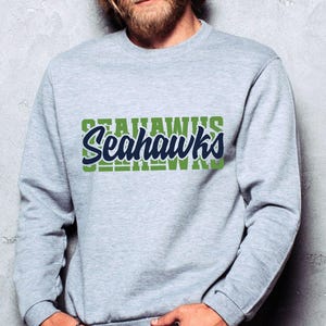 Puede incluir: Una sudadera gris claro con la palabra "Seahawks" en un dise&ntilde;o verde y azul marino. La palabra "Seahawks" est&aacute; superpuesta a un bloque verde de la misma palabra. La sudadera es de cuello redondo.