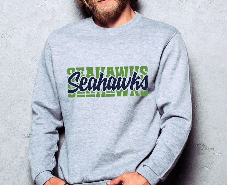 Puede incluir: Una sudadera gris claro con la palabra "Seahawks" en un dise&ntilde;o verde y azul marino. La palabra "Seahawks" est&aacute; superpuesta a un bloque verde de la misma palabra. La sudadera es de cuello redondo.
