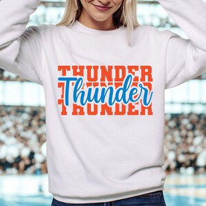May include: White sweatshirt with the word "Thunder" in a stacked design. The top word and bottom word are in a bold orange font, while the middle word is in a blue script font.