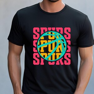 May include: Black t-shirt featuring a basketball design with the word "SPURS" in pink, stacked vertically around a teal and yellow basketball graphic. The shirt is a crew neck and appears to be made of a soft material.