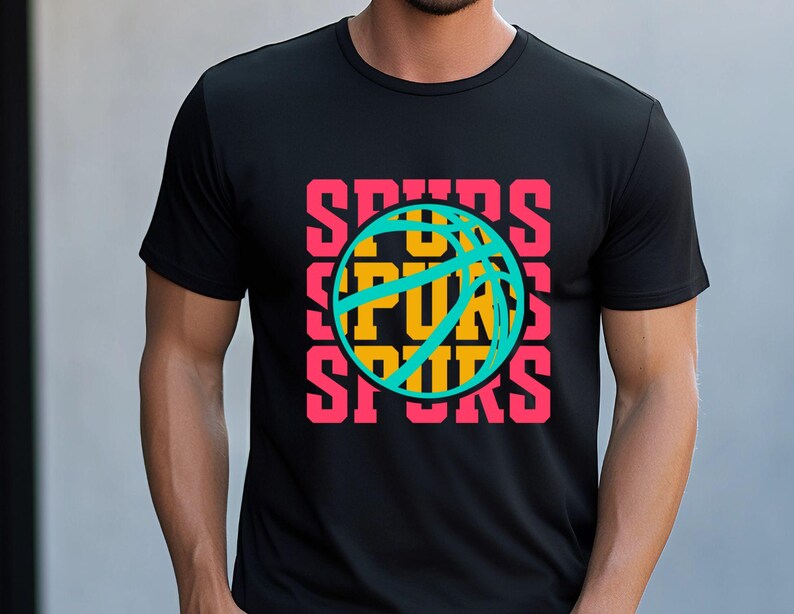 May include: Black t-shirt featuring a basketball design with the word "SPURS" in pink, stacked vertically around a teal and yellow basketball graphic. The shirt is a crew neck and appears to be made of a soft material.