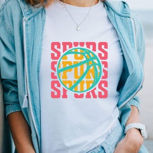 May include: White t-shirt featuring a basketball design with the word "SPURS" in pink, repeated vertically. The basketball is teal and yellow. The shirt is worn with a light blue jacket and a silver necklace.