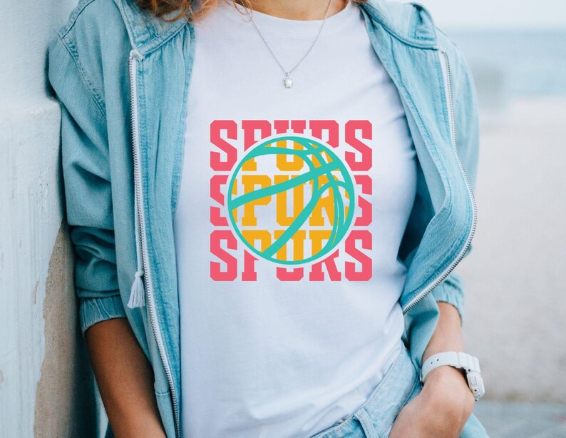 May include: White t-shirt featuring a basketball design with the word "SPURS" in pink, repeated vertically. The basketball is teal and yellow. The shirt is worn with a light blue jacket and a silver necklace.