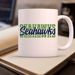 Puede incluir: Una taza de cer&aacute;mica blanca con la palabra "Seahawks" en dise&ntilde;o verde y azul. La taza tiene un asa curva y est&aacute; sobre un cuaderno con un bol&iacute;grafo.