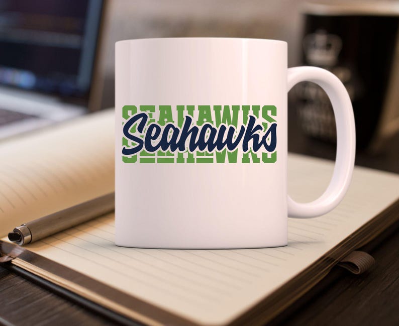 Puede incluir: Una taza de cer&aacute;mica blanca con la palabra "Seahawks" en dise&ntilde;o verde y azul. La taza tiene un asa curva y est&aacute; sobre un cuaderno con un bol&iacute;grafo.