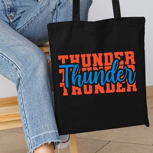 May include: Black tote bag with the word "Thunder" in orange and blue lettering. The word "Thunder" is stacked three times, with the middle word in a cursive font. The bag is held by a person wearing blue jeans.