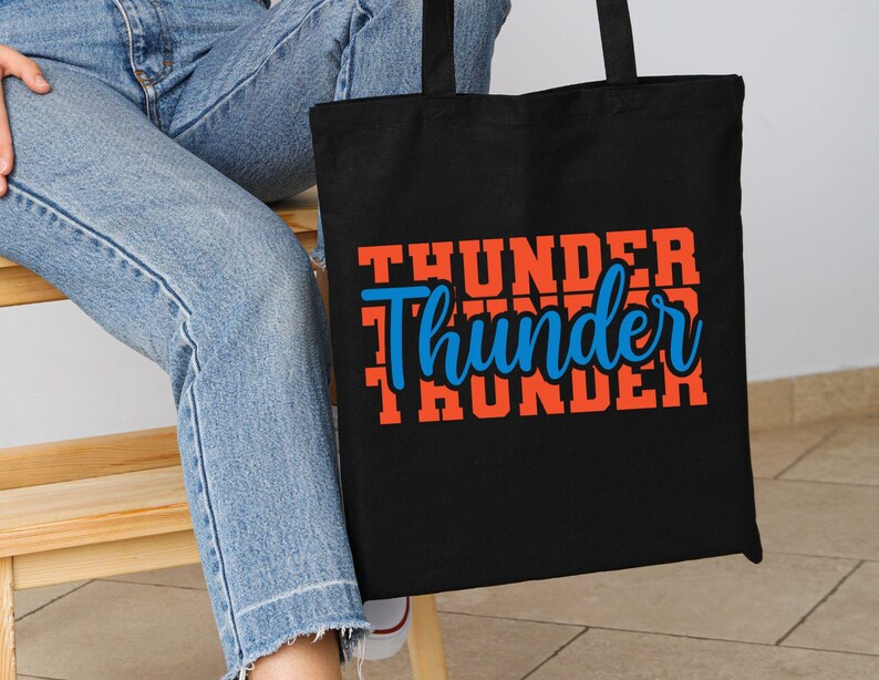 May include: Black tote bag with the word "Thunder" in orange and blue lettering. The word "Thunder" is stacked three times, with the middle word in a cursive font. The bag is held by a person wearing blue jeans.