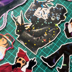 May include: A collection of holographic stickers featuring various gothic and theatrical characters. The stickers have a shiny, iridescent finish with star-shaped accents. The characters include a skeleton in a top hat and other figures in dark attire.
