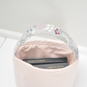 Pu&ograve; includere: Una borsa di tela rosa con un manico a fantasia floreale. La borsa &egrave; piena di due libri, "Il Libro di Mormon" e "Il Vecchio Testamento".