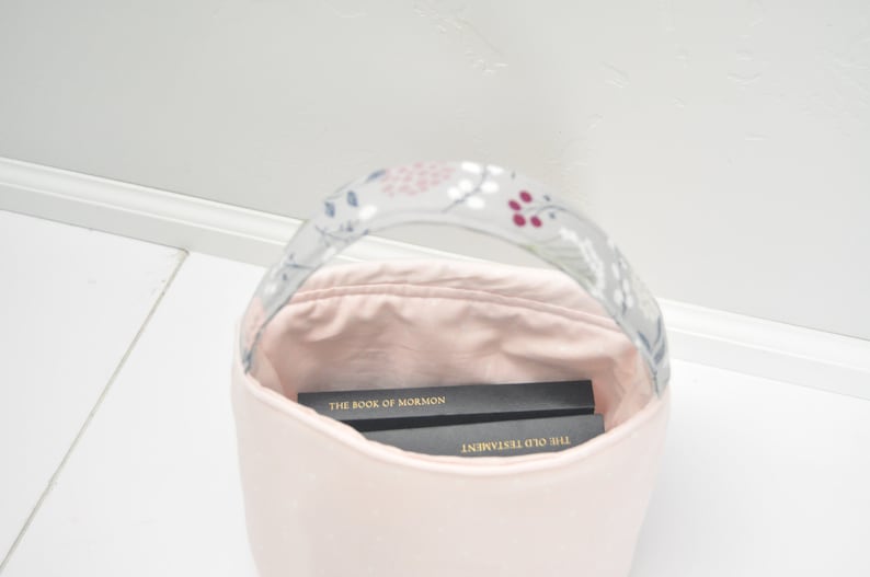 Pu&ograve; includere: Una borsa di tela rosa con un manico a fantasia floreale. La borsa &egrave; piena di due libri, "Il Libro di Mormon" e "Il Vecchio Testamento".