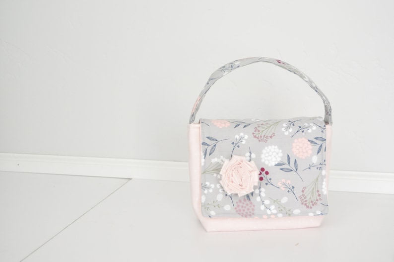 Pu&ograve; includere: Una piccola borsa a mano in tessuto grigio e rosa con stampa floreale e un accento di fiore in tessuto rosa.