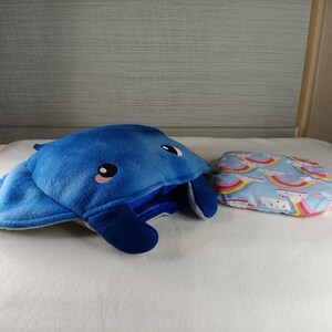 May include: A blue plush manta ray-shaped pillow with black and pink accents, next to a small rectangular pillow with a light blue background and a rainbow pattern. The manta ray pillow has a large opening.