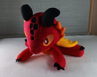 May include: A plush red dragon toy with black horns and accents. The dragon has yellow and orange wings, and black claws. The dragon's eyes are a light brown color. The toy is lying on a white surface.