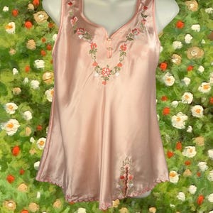 May include: A light pink satin camisole with floral embroidery around the neckline and hem. The sleeveless top has a V-neck and a scalloped hemline. The embroidery features red, green, and white flowers.