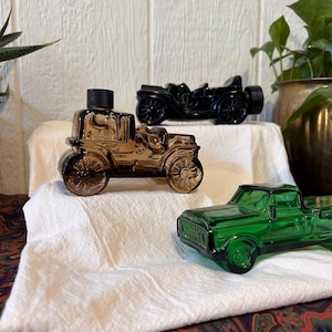 Op de afbeelding: Drie vintage auto-vormige glazen flessen in amber, zwart en groen. De amberkleurige auto heeft een zwarte dop. De zwarte auto is een klassiek model. De groene auto is een pick-up. Ze worden tentoongesteld op een witte doek.