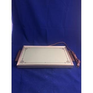 Puede incluir: Bandeja eléctrica rectangular para calentar con una superficie verde claro y detalles decorativos en las esquinas. La bandeja tiene una base de madera marrón y un cable eléctrico marrón con un enchufe de dos clavijas. La bandeja está diseñada para mantener la comida caliente.