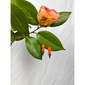 May include: A pair of orange and silver earrings with a geometric design. The earrings are displayed on a green leafy branch with a peach-colored flower against a white background. The earrings have a modern, minimalist style.