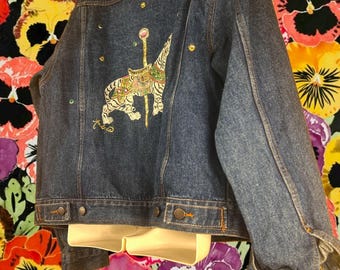 Könnte beinhalten: Dunkelblaue Jeansjacke mit einem farbenfrohen, gestickten Tigerdesign auf dem Rücken. Die Jacke hat goldfarbene Knöpfe und dekorative Verzierungen. Ein einzigartiges Oberbekleidungsstück.